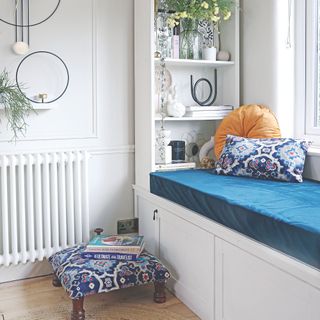 White corner of a room with a blue cushioned bench seat, a patterned foot stool on the floor, and a bookshelf behind