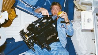 a man in a blue jacket works with a large black camera on board nasa&#039;s space shuttle