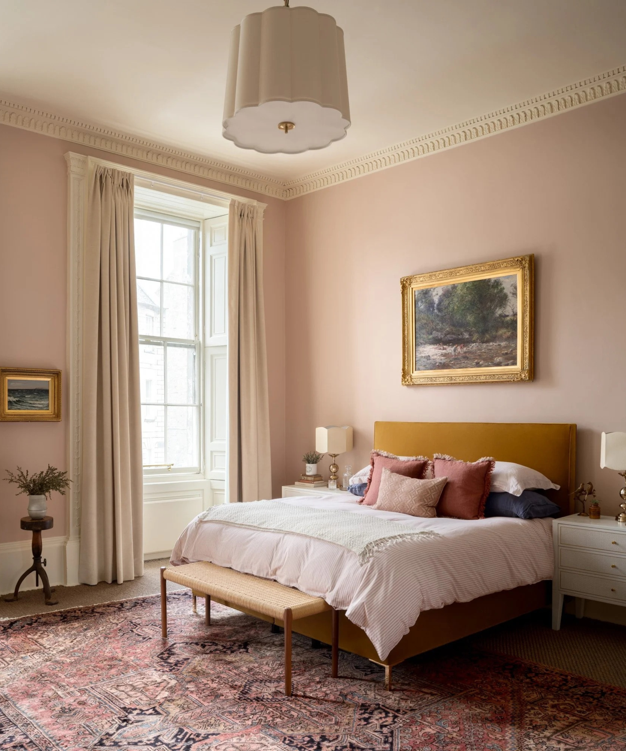 a bedroom with high ceilings, pink painted walls, a large window, a mustard-colored bed frame, a bed with several pillows on top, and a Persian rug on the floor