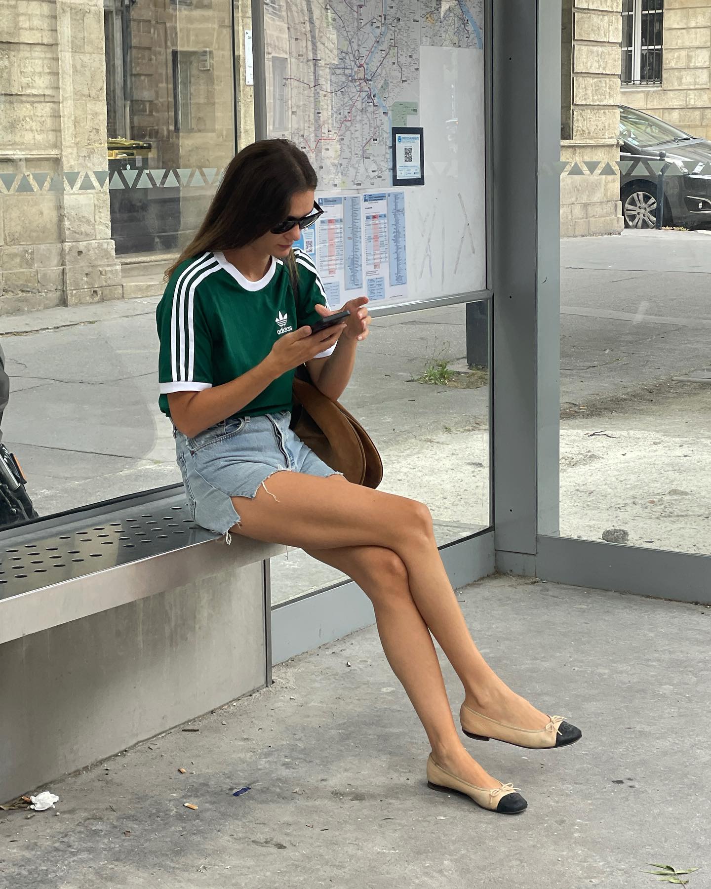 French fashion influencer Anne Laure Mais sits on a bench at a bus stop in Paris wearing a green striped Adidas t-shirt, cut-off denim shorts, and Chanel cap-toe flats
