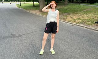 Vasilisa Kirilochkina pauses for a photograph ona run; she is in a vest and running shorts, standing in the middle of a park roadway, smiling at the camera. Her hand rests on her brow as she shades her eyes from the sun.