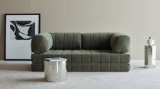 A green piano key style sofa in a minimalist living room with neutral walls and carpet and chrome side tables and a framed artwork leaning against the wall on the floor behind