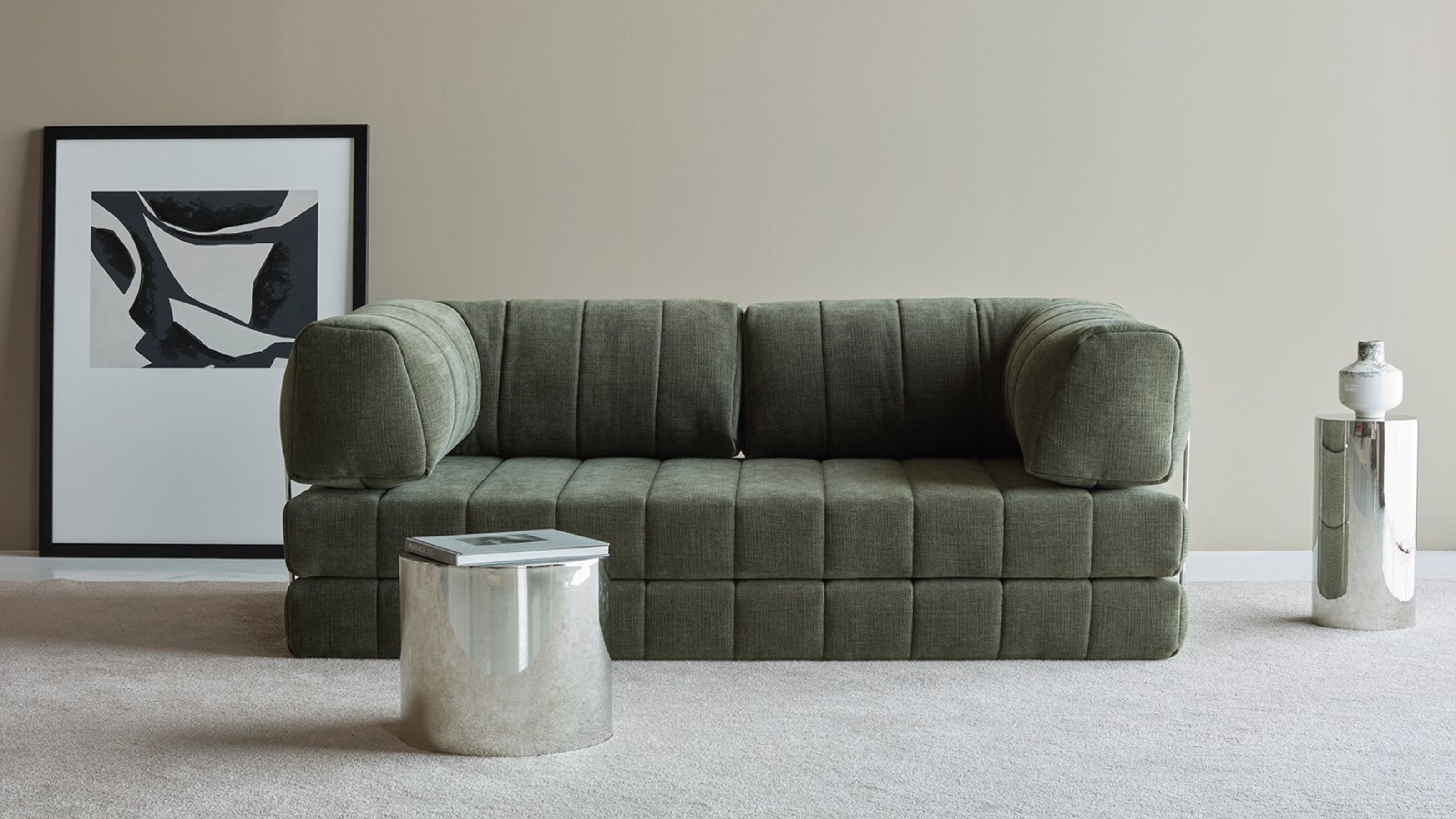 A green piano key style sofa in a minimalist living room with neutral walls and carpet and chrome side tables and a framed artwork leaning against the wall on the floor behind