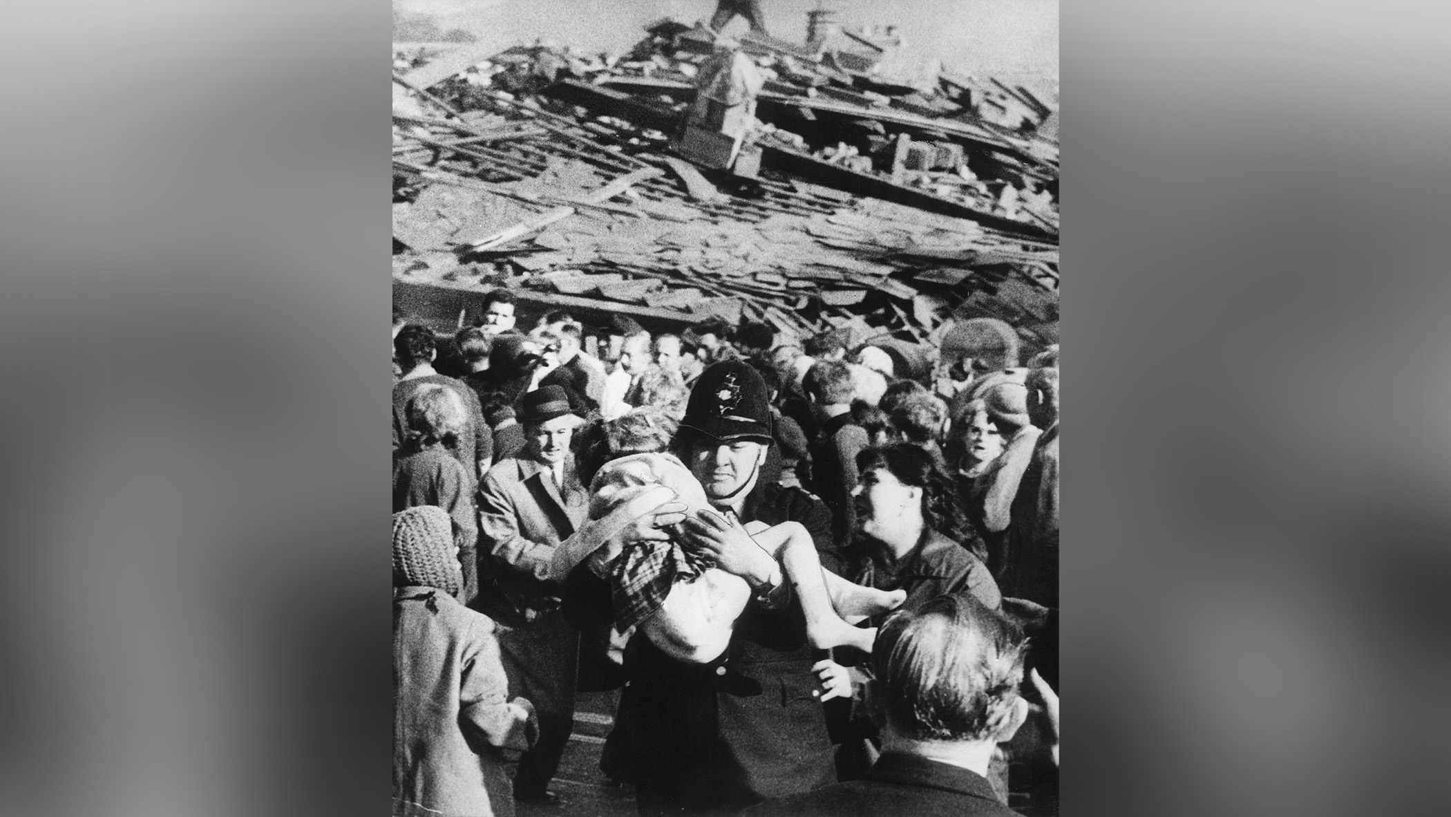 Police officer carries limb child as woman looks on among crowd of people digging in background.