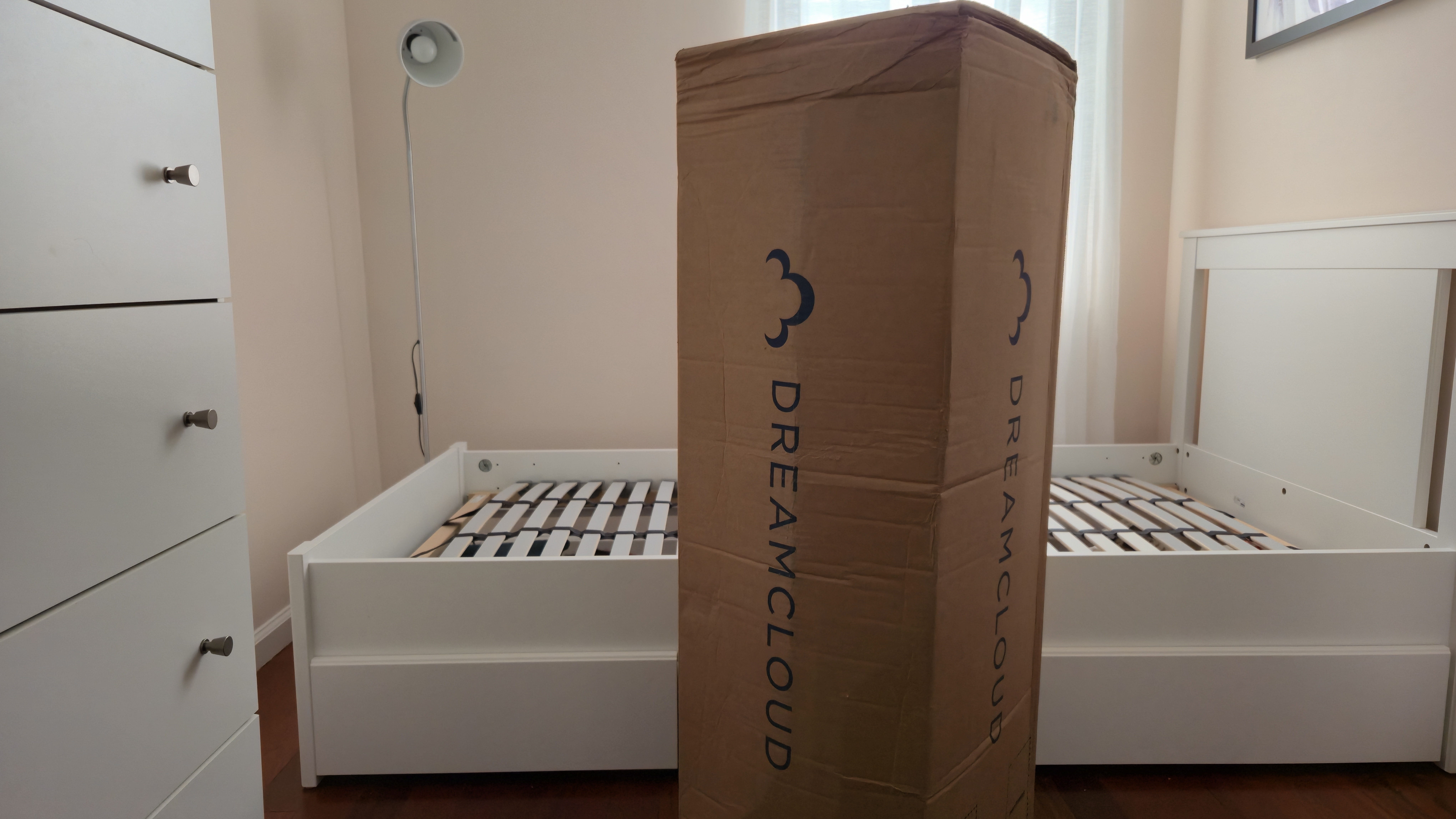DreamCloud Classic Memory Foam Mattress, standing upright in its box next to a white platform bed