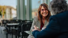 An attractive older woman in business casual smiles at a man sitting opposite her at an outdoor cafe table. They may be having a meeting or a friendly discussion.