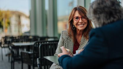 An attractive older woman in business casual smiles at a man sitting opposite her at an outdoor cafe table. They may be having a meeting or a friendly discussion.