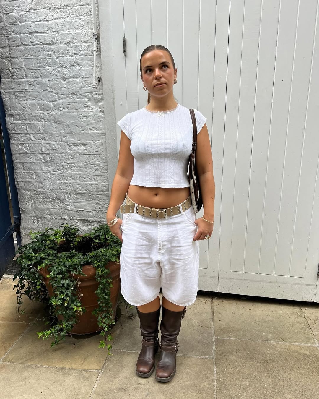 A woman standing wearing knee-length white shorts, a belt, a white tee, and brown leather motor boots.