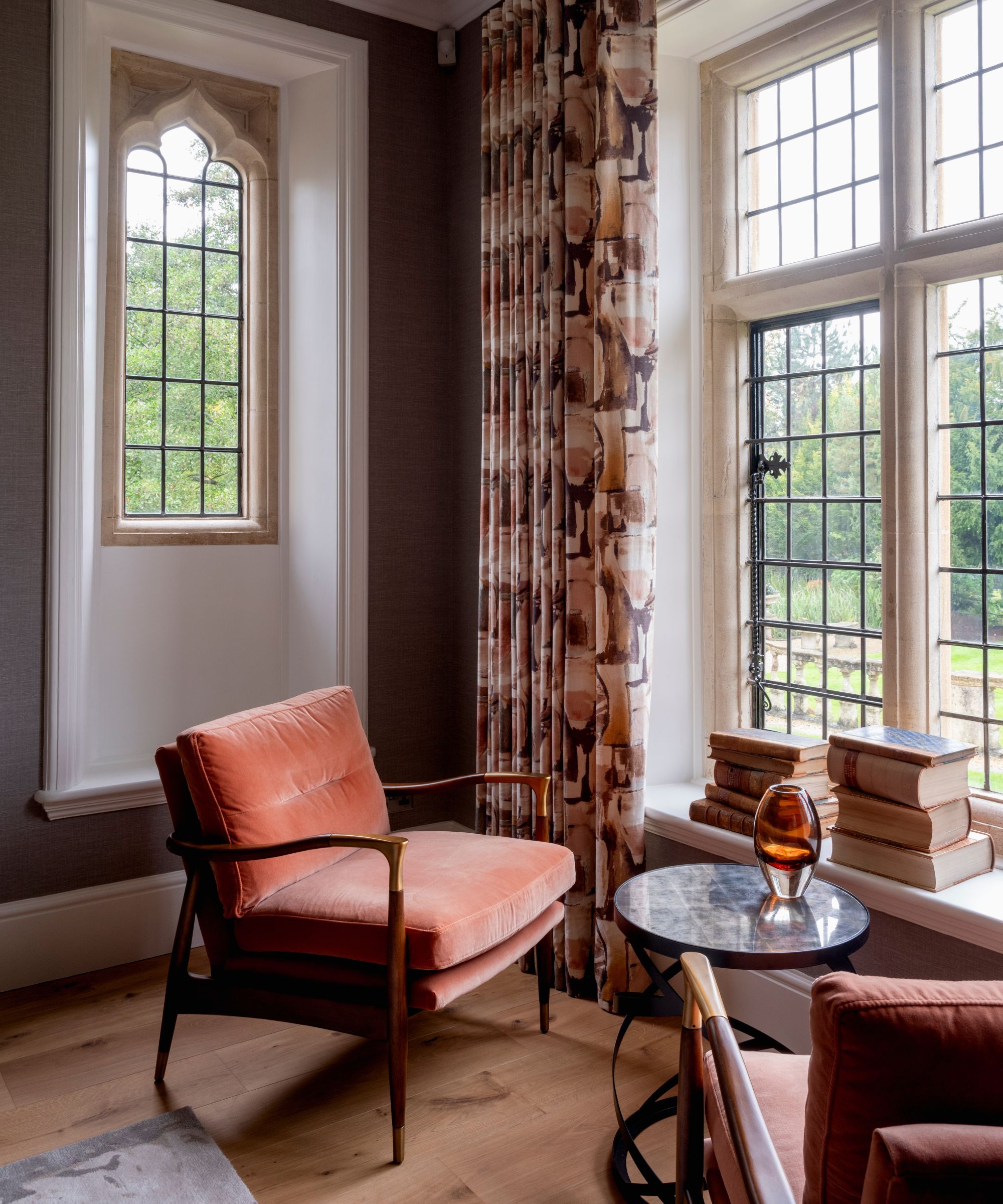 A terracotta velvet mid-century armchair beside tall leaded Gothic windows in a moody taupe room, with abstract print curtains, stacked books on the sill and a marble side table.