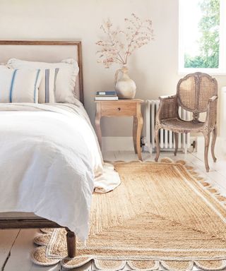A scalloped edge jute rug on the floor beside of a white bed in a bright bedroom