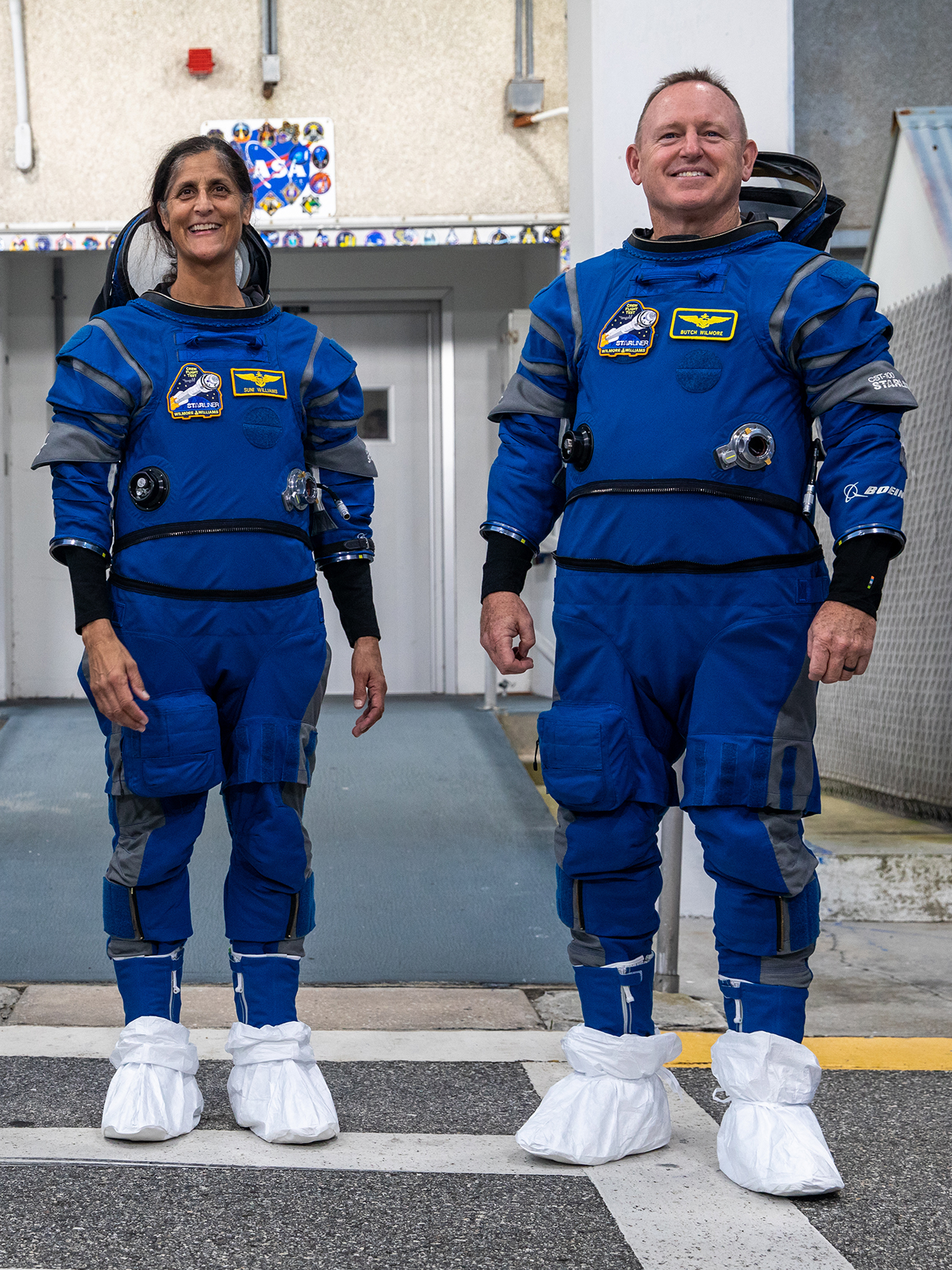two astronauts standing side by side on a tarmac in spacesuits.  white covers protect their boots