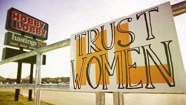 'Trust Women' sign posted on barrier in front of 'Hobby Lobby' and 'Hastings' signs
