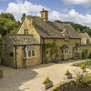 Cotswold stone cottage with paved driveway, trees and garden and blue skies