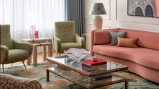 Living room with white walls, coral sofa, green armchairs, green and coral rug, glass coffee table and pink chandelier