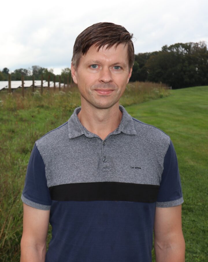 A man with short hair wearing a blue and gray t-shirt looks at the camera.
