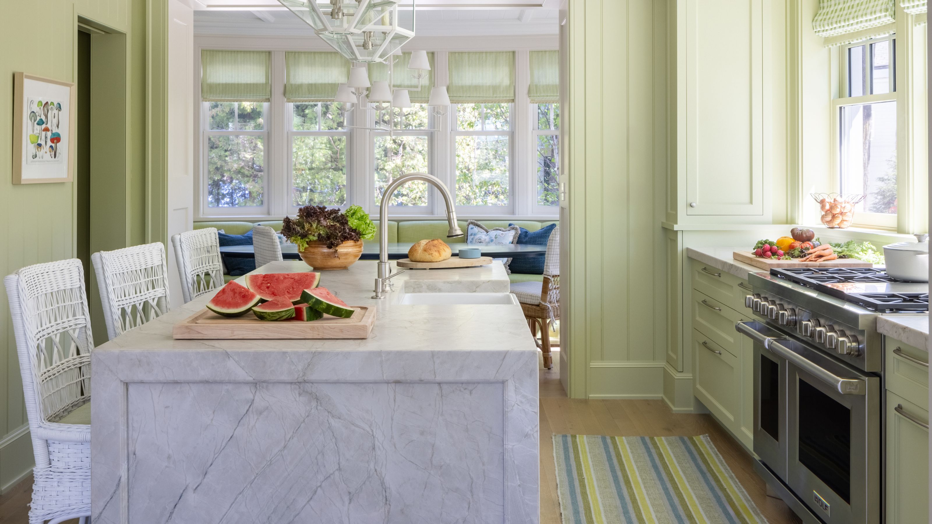 a pale green kitchen with a large marble island, striped rug, and a archway through to a banquette dining area