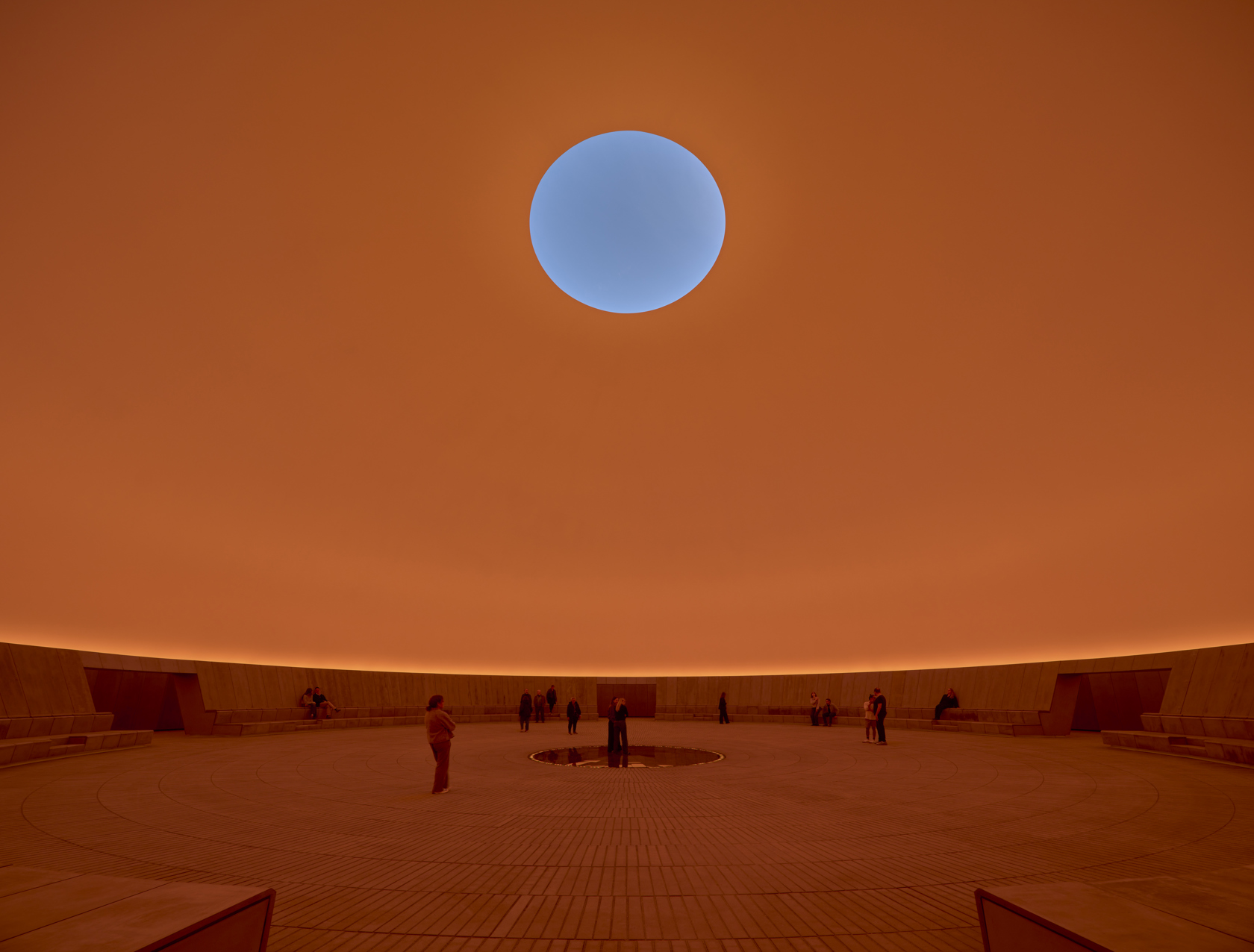 An orange-tinted, semi-empty spherical theater with a ceiling porthole window after which one can see a blue sky.