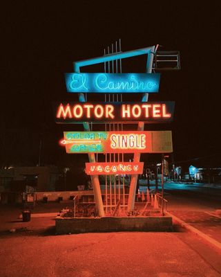 A road sign features neons reading, from top to bottom, "El Camino", "MOTOR HOTEL", "Color TV Office", "SINGLE", and "VACANCY", bringing light to nighttime.