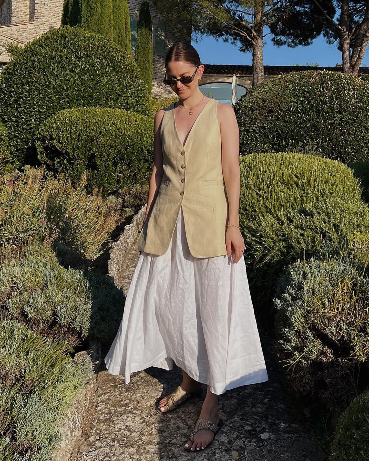Influencer Marissa Cox poses in a garden in Southern France wearing oval sunglasses, a neutral vest top, white linen skirt, and snakeskin flat sandals.