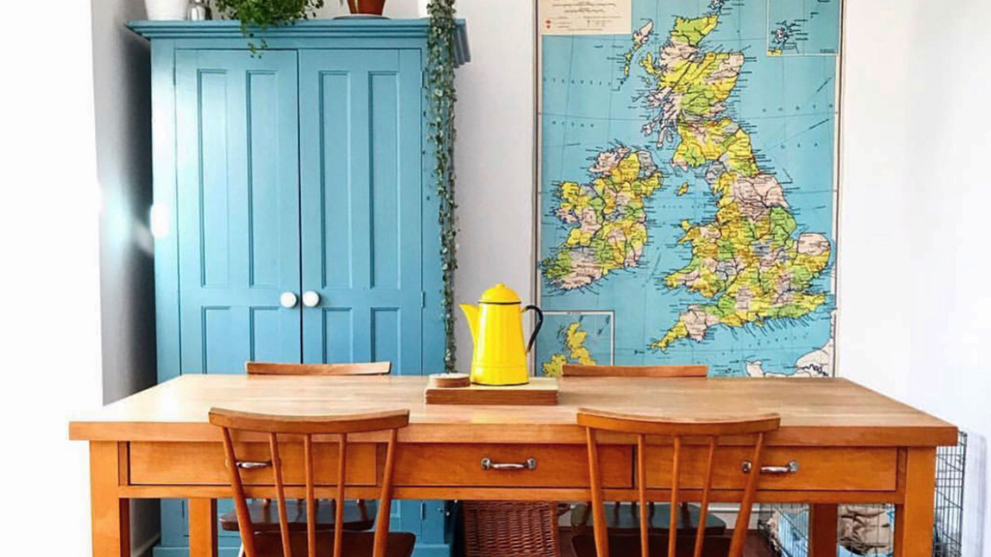 Kitchen with wooden table and chairs in front of blue painted cabinet topped with houseplants beside vintage map of Britain and Ireland hanging on wall