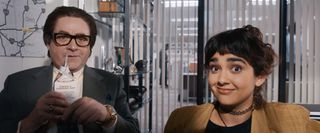 Ty (Zach Galifianakis) and Maya (Geraldine Viswanathan) in The Beanie Bubble. The shot is taken from the perspective of inside a computer monitor as the two of them gaze into it. Ty is smiling and drinking a carton of chocolate milk through a straw, and Maya is smiling widely in disbelief