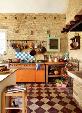 a rustic freestanding kitchen, with a giant orange aga, lots of freestanding units storing food and pots and pans, and a hanging rack full of pots and pans - with a purple and white tiled floor and exposed brick walls