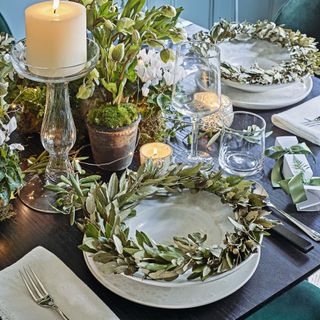 A place setting for Christmas with white candles, glass candlestick, silver green leaf wreaths and pots of greenery, ivy, and moss.