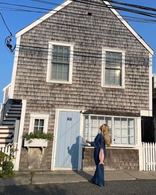 Influencer Martine Lavelle poses in front of a classic Cape Cod home with a light blue door wearing a blue Realisation maxi dress and fish-shaped bag.