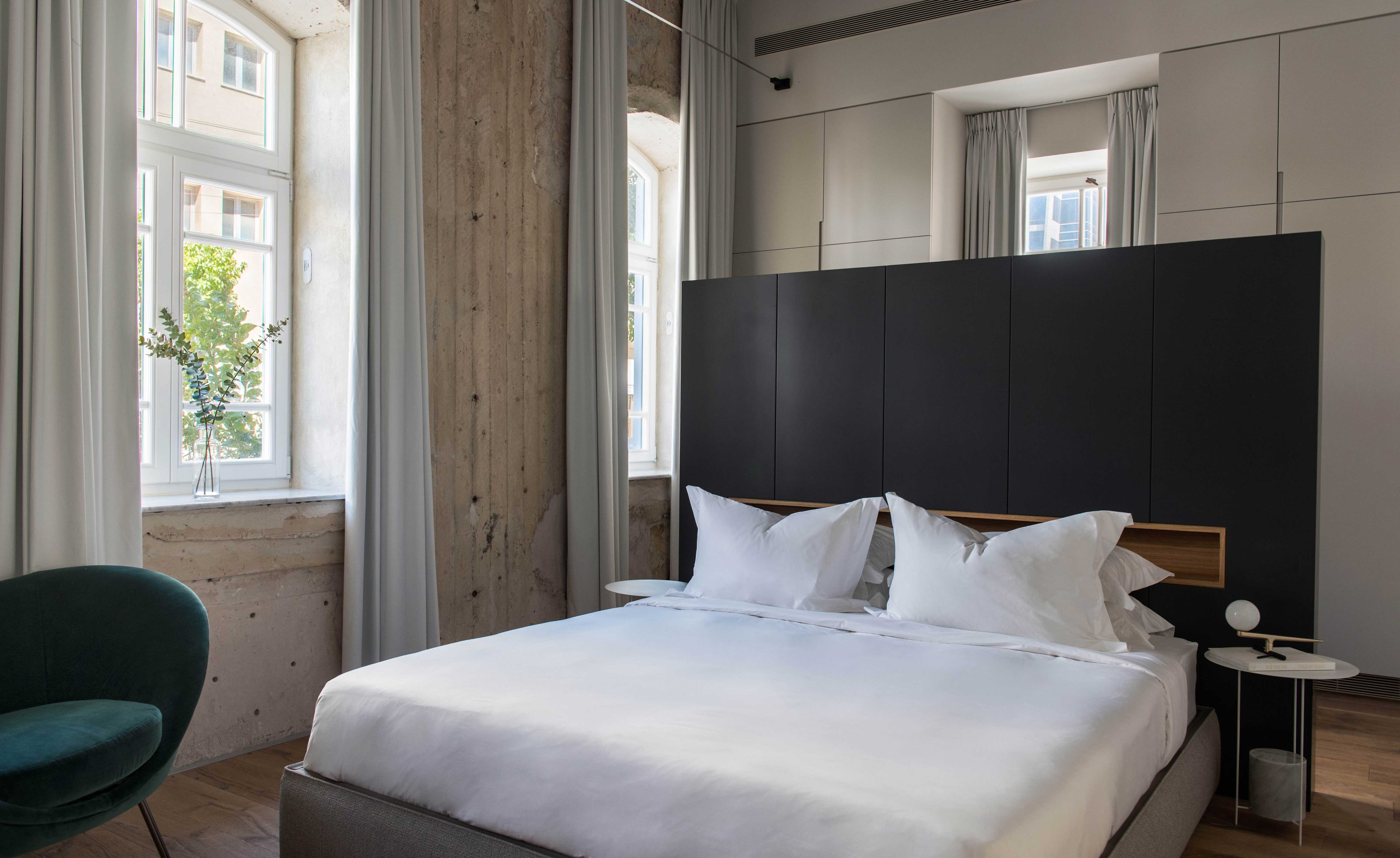 Bed with dark headboard, exposed wall finish, single designer chair with high hanging curtains