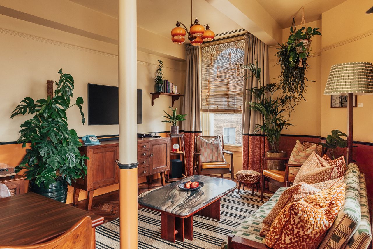 A layered, transitional hotel suite featuring a series of figurative, colorful artworks on its walls, patterned furniture, including rugs, armchairs, and sofas, split-color pillars, and a buttery, tile blue, and green palette.