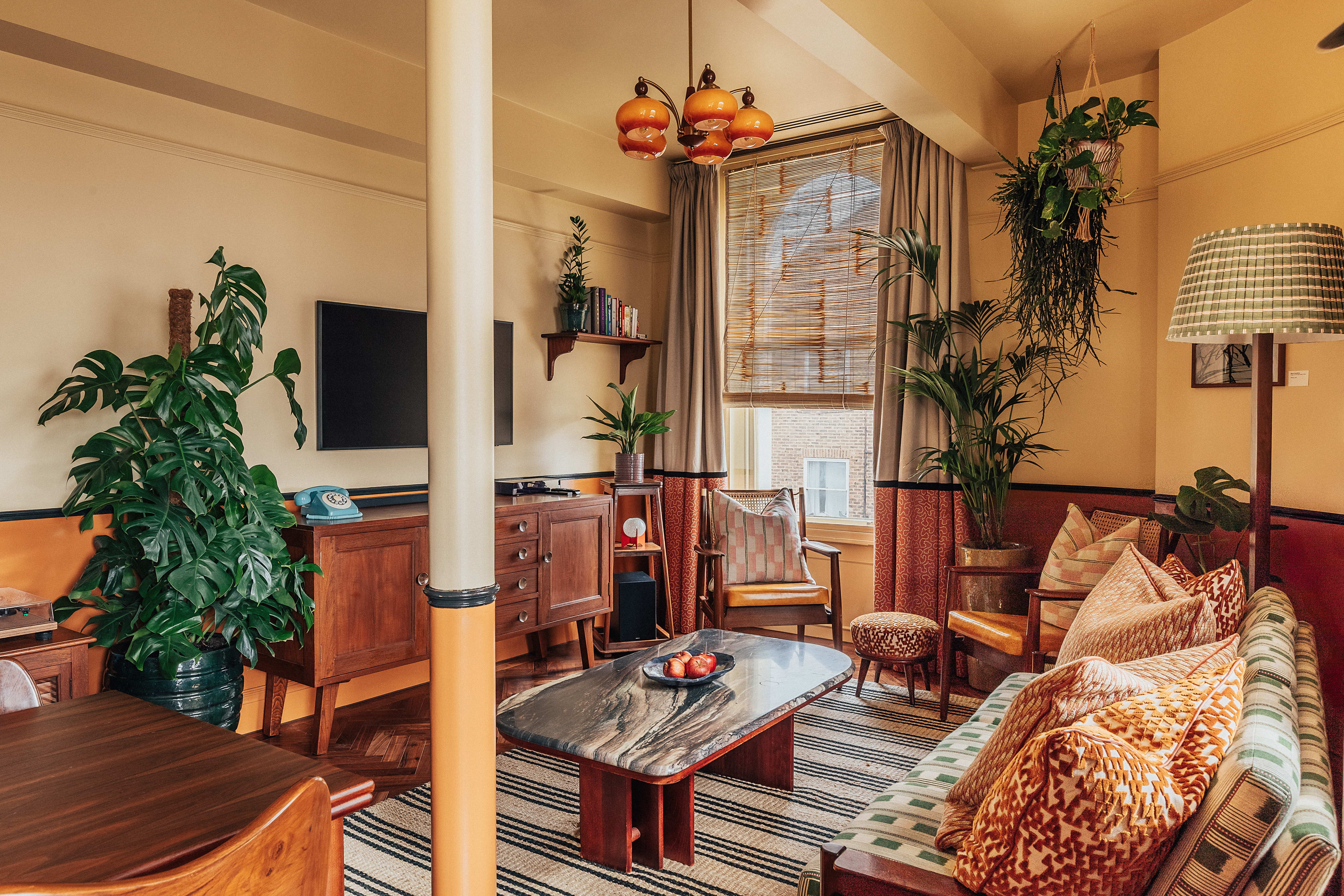 A layered, transitional hotel suite featuring a series of figurative, colorful artworks on its walls, patterned furniture, including rugs, armchairs, and sofas, split-color pillars, and a buttery, tile blue, and green palette.