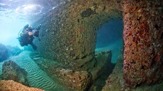 Scuba diver swimming through submerged ancient Roman building ruins with a big still intact stone arch.