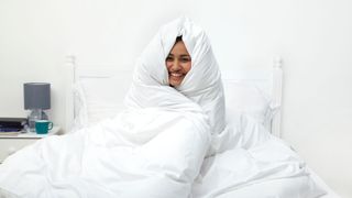 A woman wrapped up in a white duvet sat on her bed smiling and laughing
