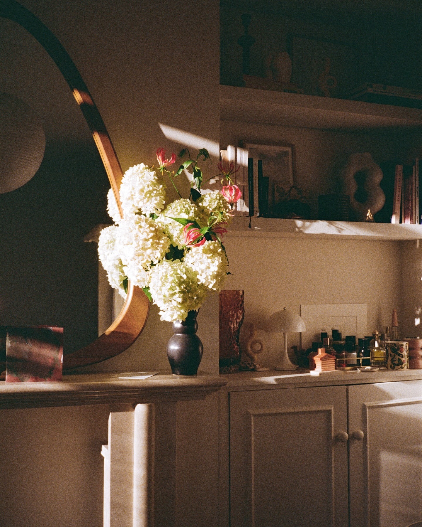 An evening sun dappled living room with mantlepiece, a wooden mirror and shelving with beauty products