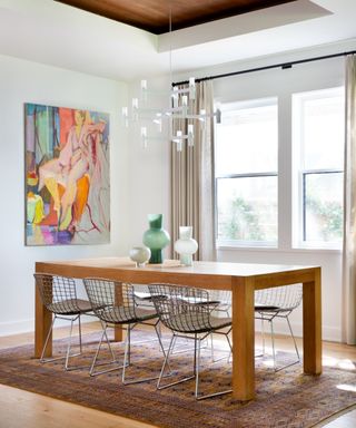 A dining room with a large wooden table, basket metal chairs, abstract art and a contemporary candle chandelier