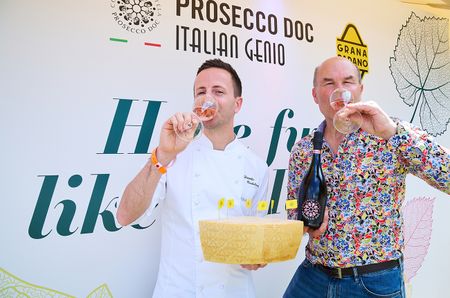Two men drinking glasses of Prosecco ros&eacute;, one in a chef's uniform, holding a large chunk of Grana Padano