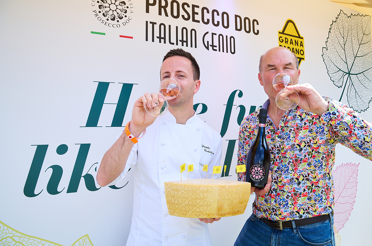 Two men drinking glasses of Prosecco ros&eacute;, one in a chef's uniform, holding a large chunk of Grana Padano