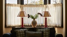 Two lamps sit beside a window and in the middle of the two lamps is a vase with flowers inside. Below the lamps is a sofa with cushions on top.