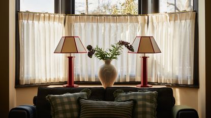 Two lamps sit beside a window and in the middle of the two lamps is a vase with flowers inside. Below the lamps is a sofa with cushions on top.