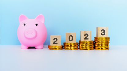 A pink piggy bank sits next to four piles of coins that get progressively bigger with the year 2023 spelled out in blocks on top.