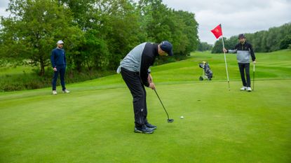 Neil Tappin hitting a putt on the green, watched by Barry Plummer and Dan Parker, at Burhill Golf Club