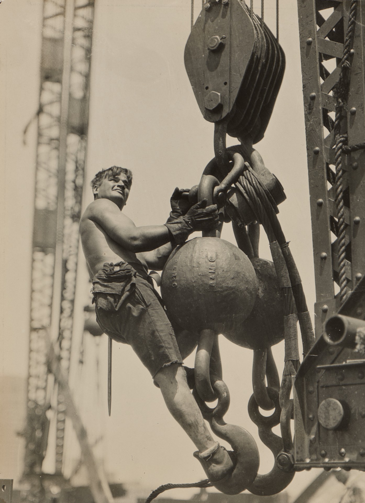 A shirtless construction worker grins as he clings with easy confidence to a giant iron hook and pulley high above the Empire State Building construction site.