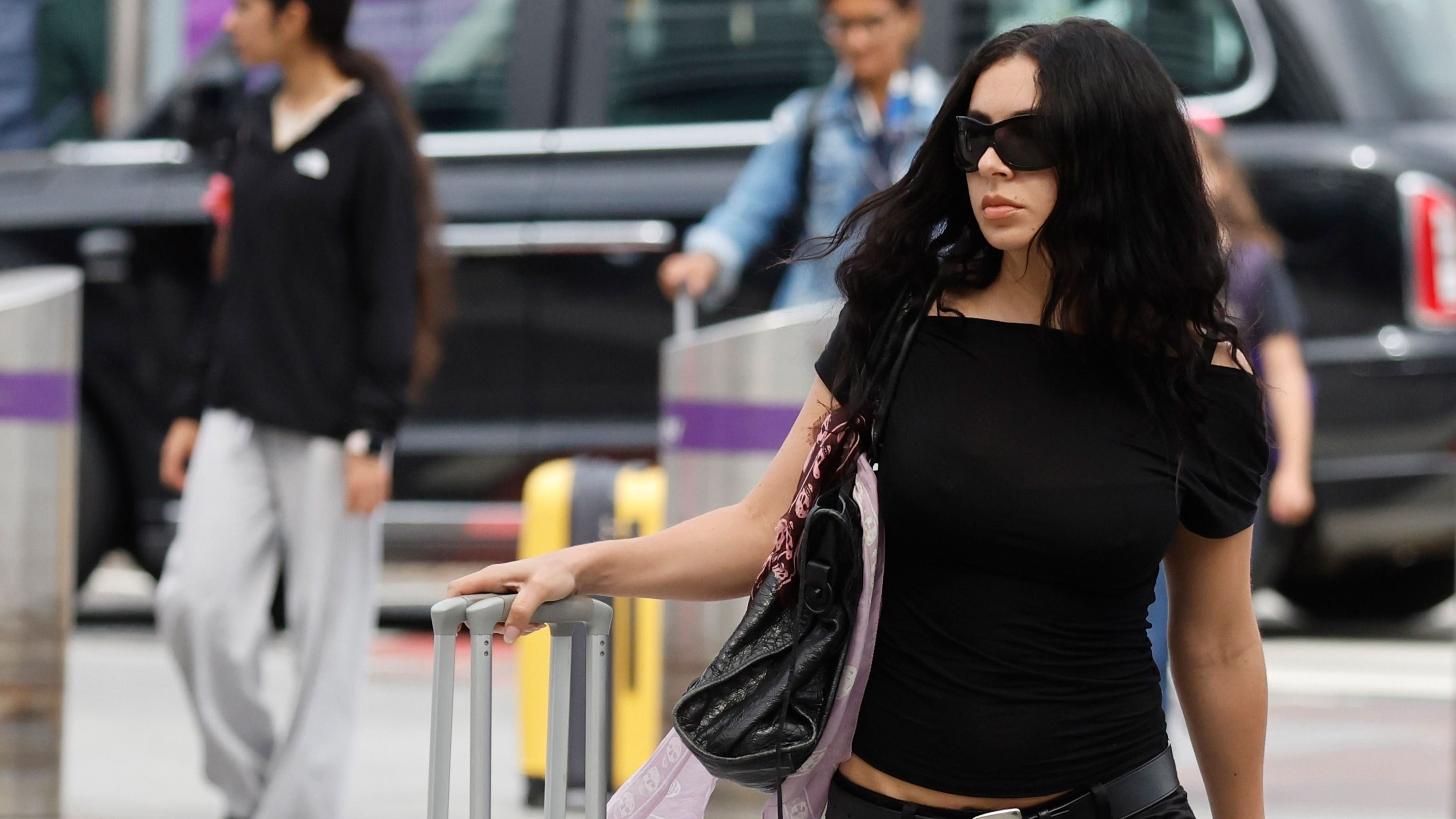 Charli XCX at Heathrow airport carrying aluminum luggage, wearing a black top, black pedal pushers, and Maison Margiela Tabi flats.