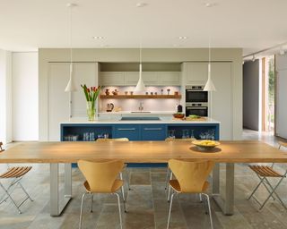 An example of modern kitchen lighting ideas showing a long wooden dining table in front of a blue island with white pendant lights above