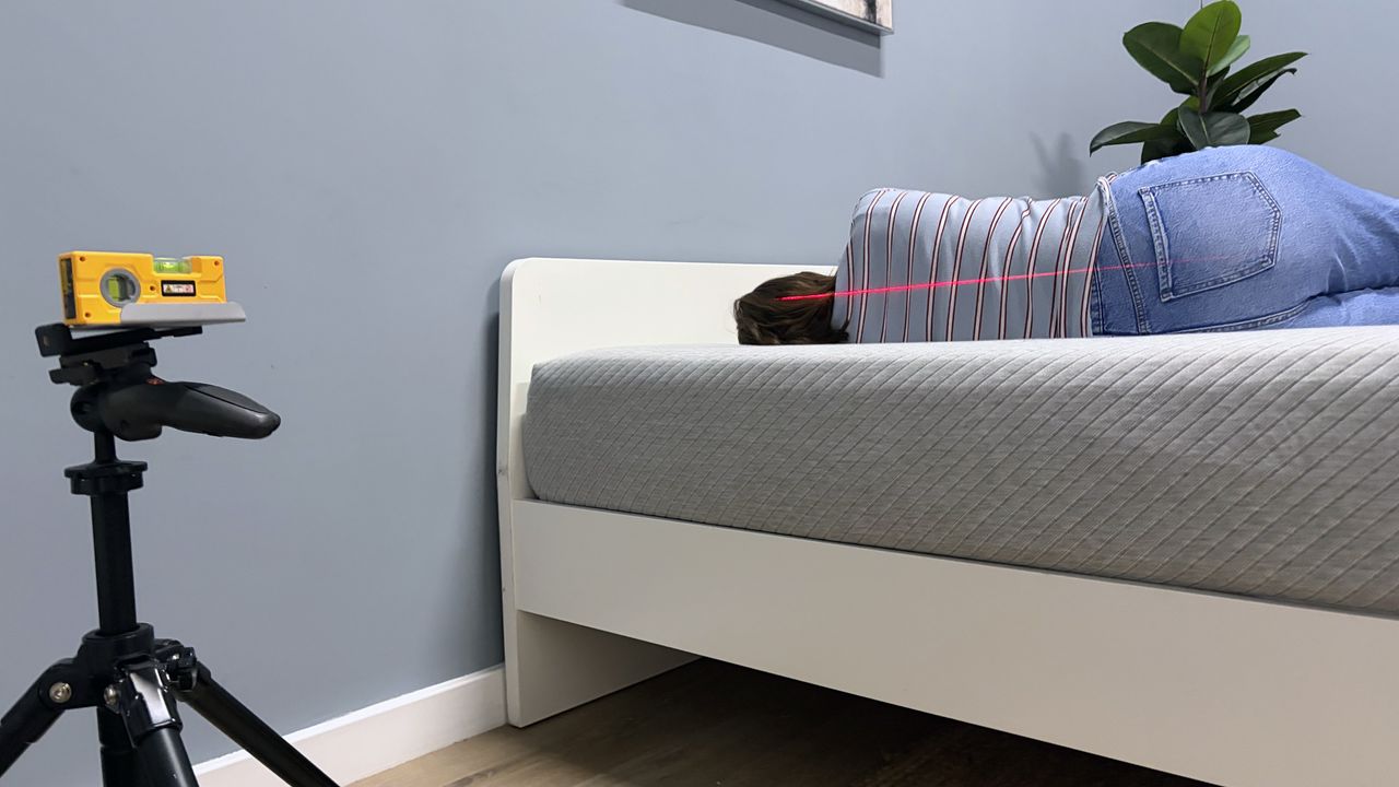 Tester Ruth Jones lies on the Leesa Original mattress in the side sleeping position. A red laser line can be seen across her back. On the left hand side of the screen is a laser measure balanced on a tripod.