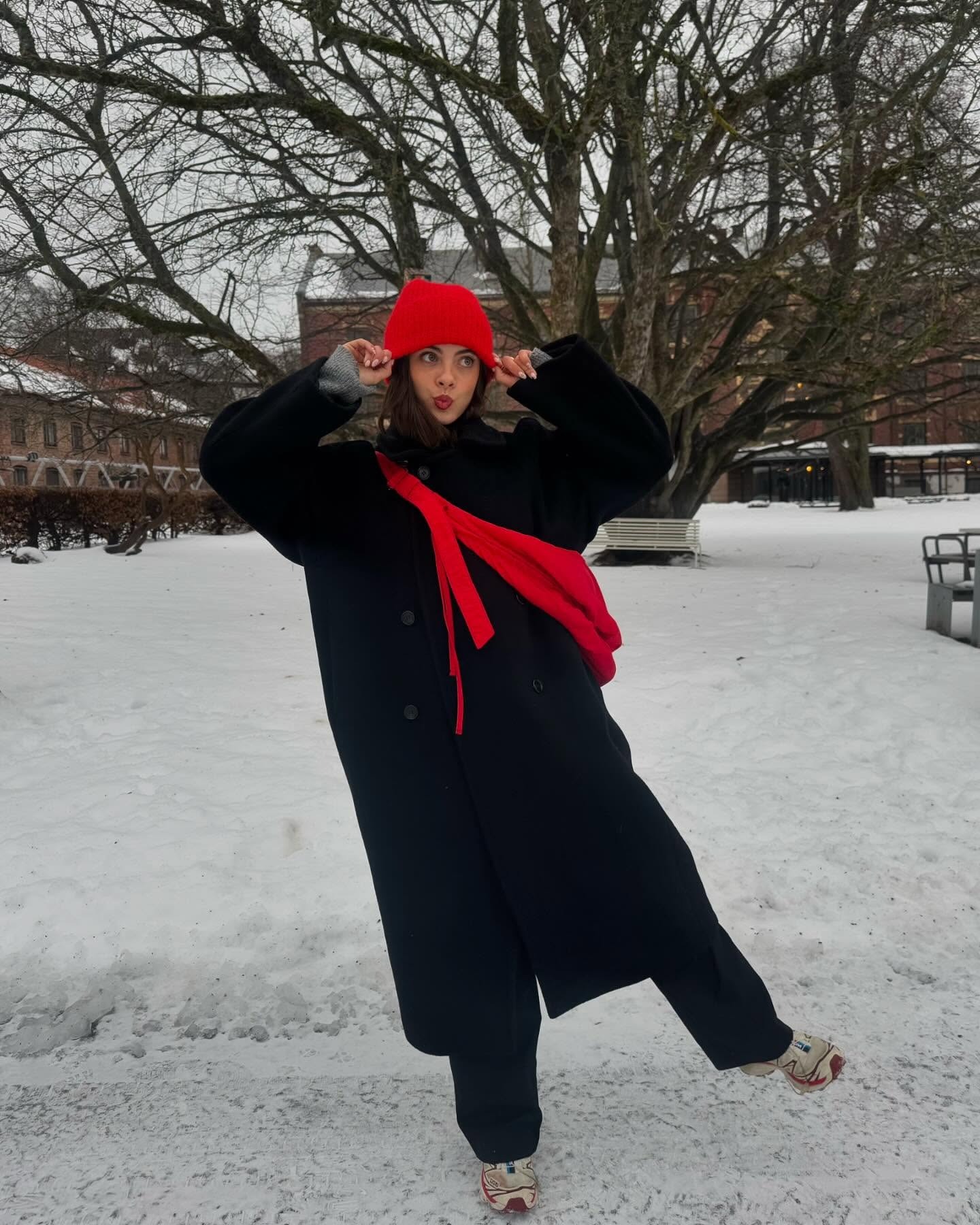 Influencer @tia_dewitt wears a black coat with a red beanie and red knit over her shoulder in the snow.