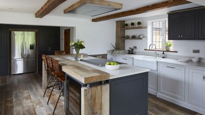 A kitchen with blue-grey island with reclaimed wood-topped breakfast bar, exposed wooden beams on a white ceiling, and white quartz worktops over pale grey cabinets