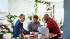 A financial adviser sits with a smiling couple at their kitchen table and shows them financial figures in paperwork.