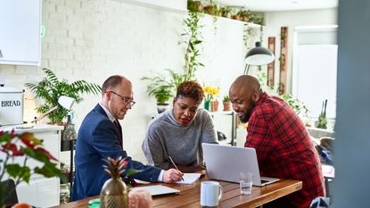 A financial adviser sits with a smiling couple at their kitchen table and shows them financial figures in paperwork.