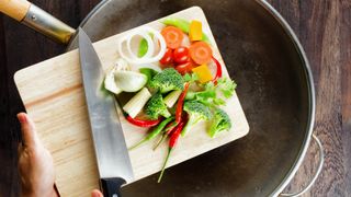 Vegetables being scraped from a cutting board into a pan with a knife